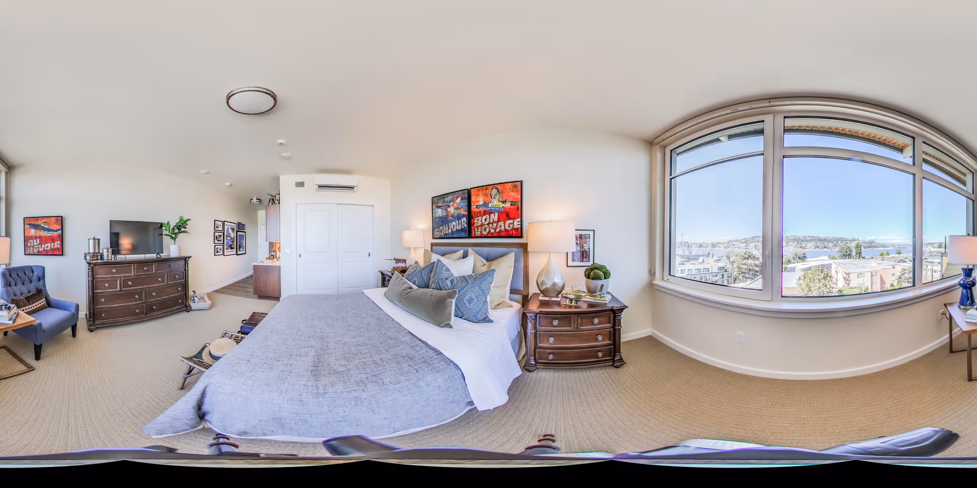 A bright and spacious bedroom in a senior living facility featuring a large bed with multiple pillows, two bedside tables with lamps, a dresser with a TV on top, a blue armchair, and large windows offering a scenic view of the outdoors including buildings and water in the distance.