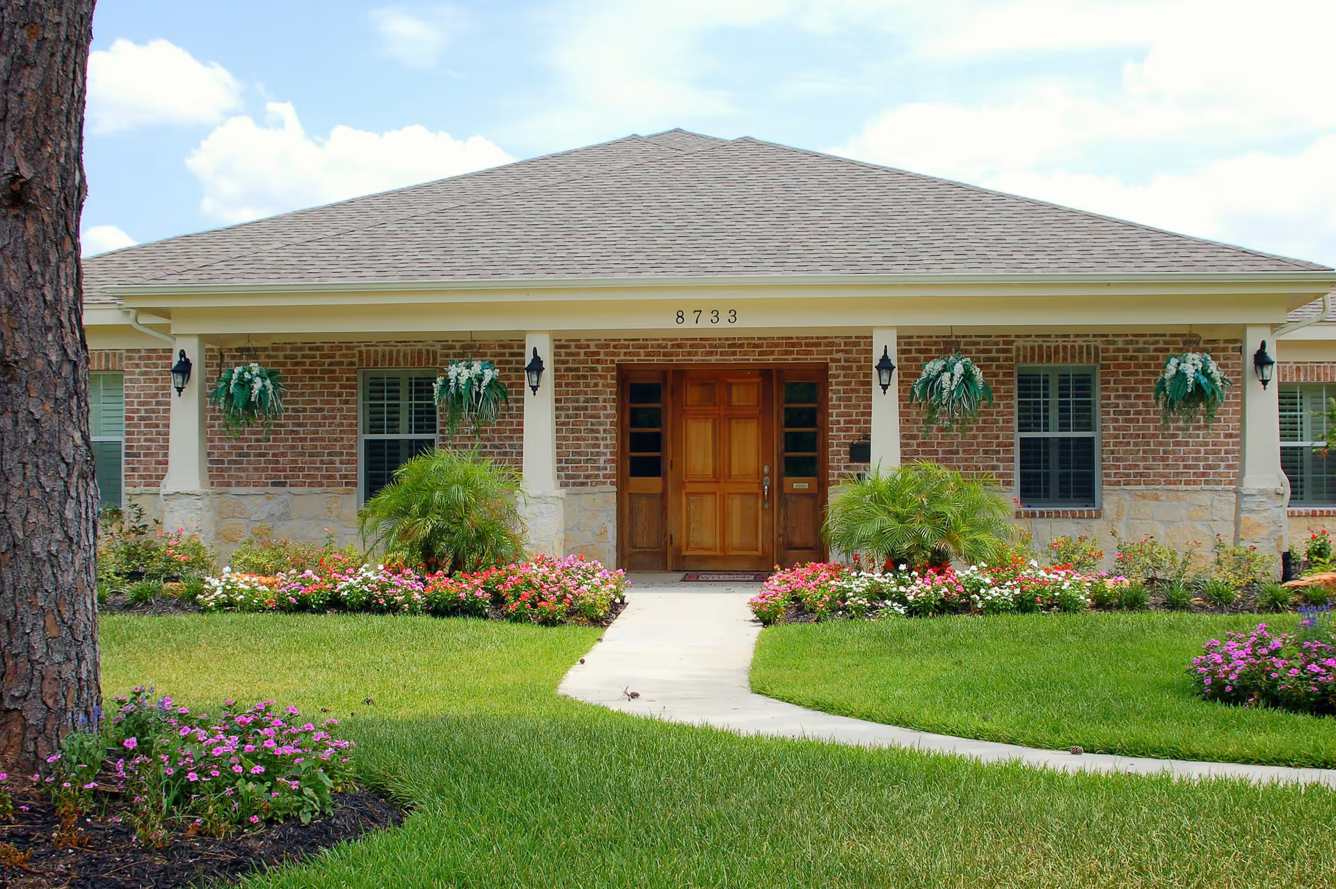 Front exterior view of a single-story brick building with a wooden door, four columns supporting a covered porch, hanging flower baskets, and well-maintained flower beds and green lawn in front.