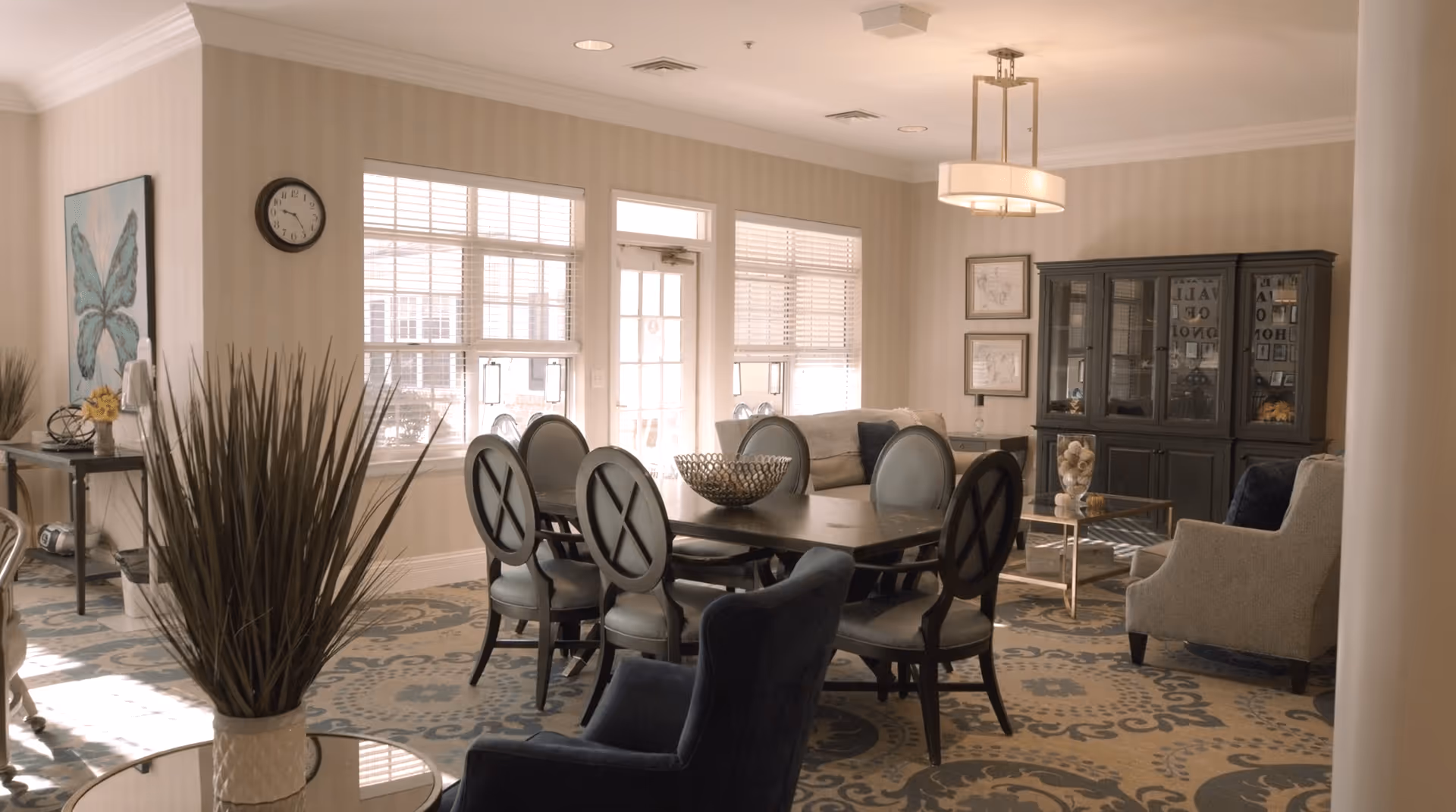 A well-lit living room and dining area in a senior living facility featuring a dark wooden dining table with six chairs, a beige sofa, armchairs, a glass-front cabinet, and decorative wall art. Large windows with blinds allow natural light to fill the space, and a patterned carpet covers the floor.