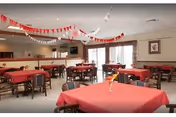 A dining room with multiple tables covered in red tablecloths and surrounded by wooden chairs. The room is decorated with red and white hanging streamers. There is a window with curtains on one side and a television mounted on the wall in the background.