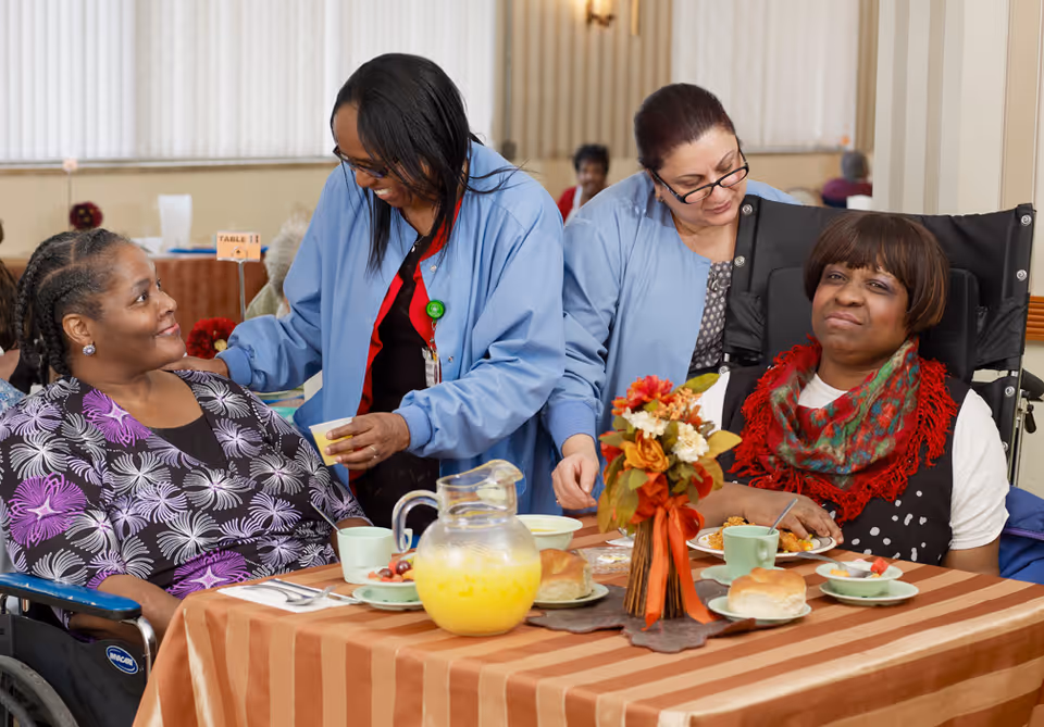 Two elderly women seated at a dining table with plates of food and a pitcher of orange juice, accompanied by two caregivers in blue uniforms attending to them in a communal dining area.
