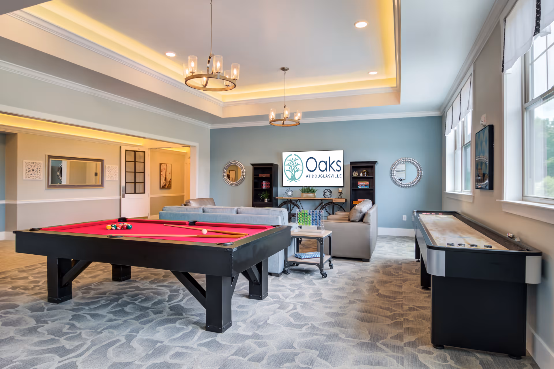 A bright and spacious lounge area in Oaks at Douglasville featuring a red felt pool table, a shuffleboard table, and comfortable seating with sofas and a coffee table. The room has large windows letting in natural light, two modern chandeliers, and a TV mounted on a blue accent wall displaying the Oaks at Douglasville logo. Decorative mirrors and shelves with plants and books add to the cozy atmosphere.