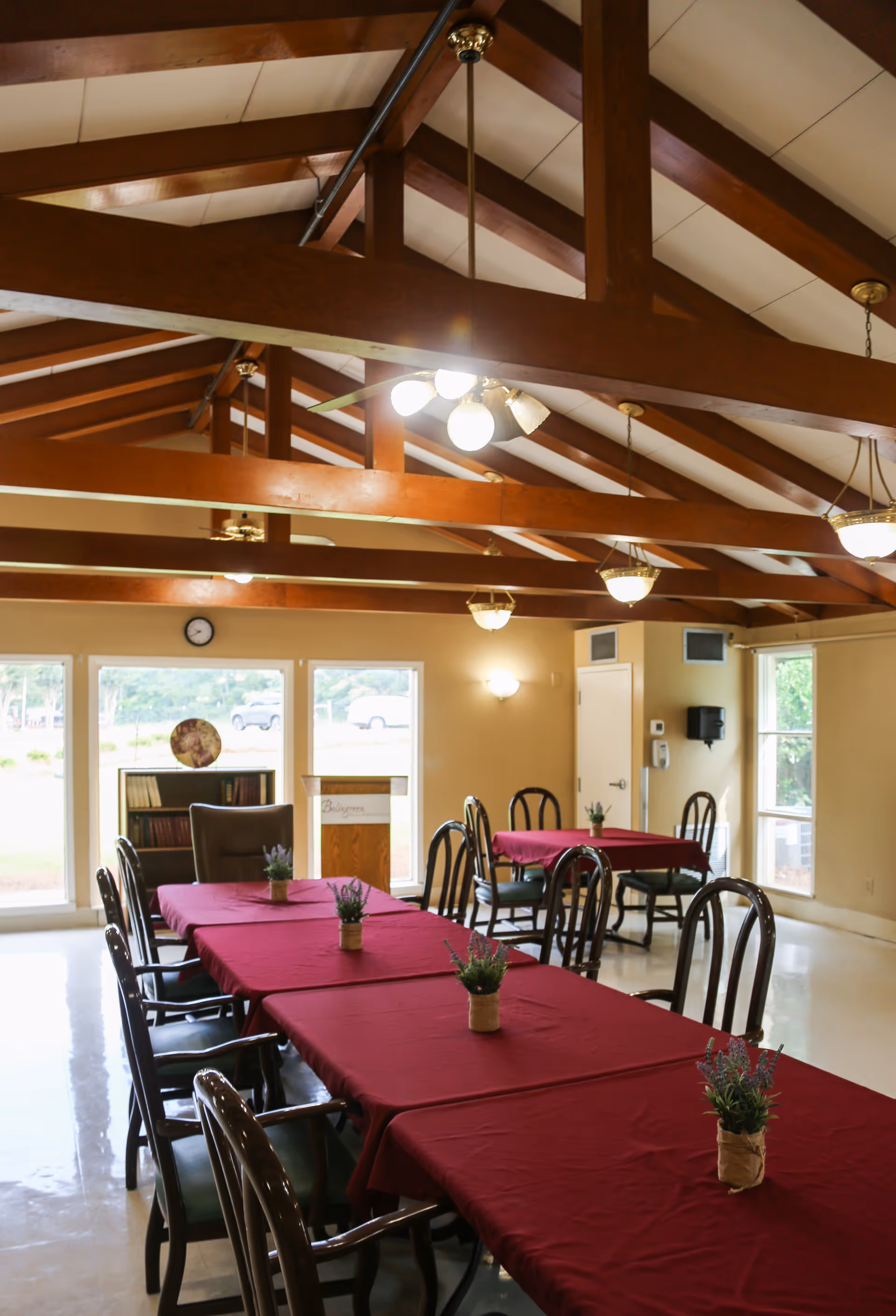 Communal dining room with long tables covered in red tablecloths, wooden chairs, small potted plants, and exposed wooden ceiling beams.