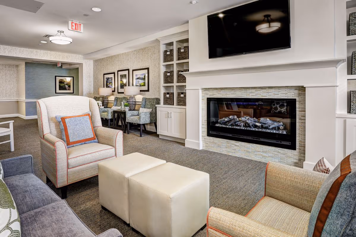 A cozy living room area in a senior living facility featuring a modern electric fireplace with a mounted flat-screen TV above it. The room includes comfortable armchairs with accent pillows, a gray sofa, two cream-colored ottomans, and a row of patterned chairs with side tables and lamps against the wall. The walls are decorated with framed artwork and built-in shelves with storage baskets. An exit sign is visible on the ceiling.