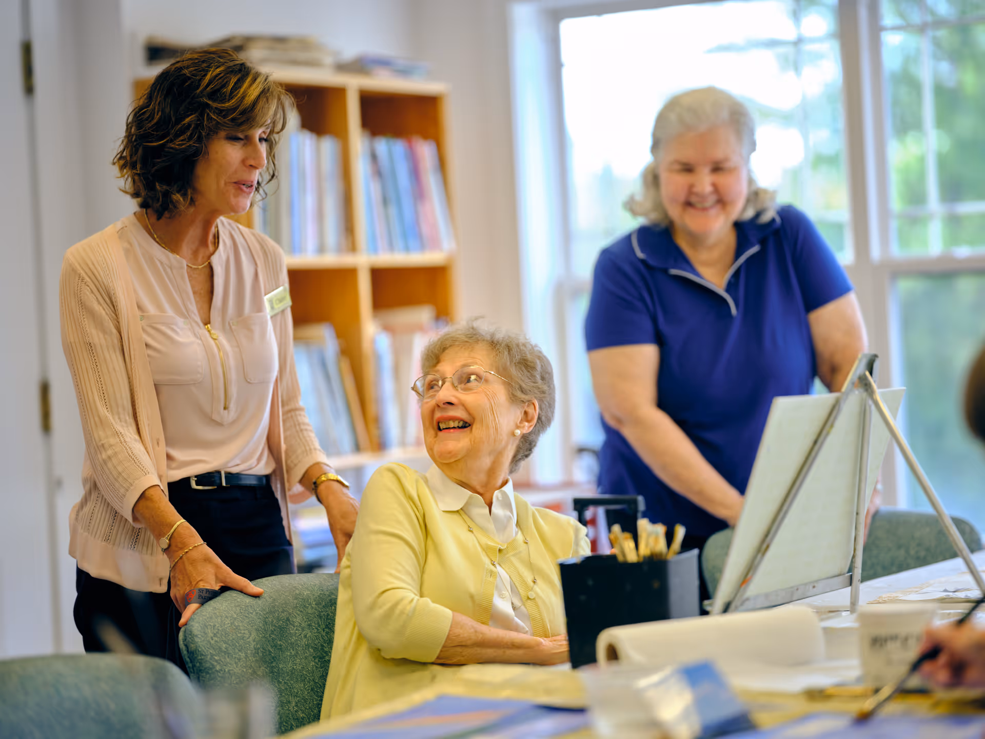 Three elderly women in a well-lit room with large windows. One woman in a yellow cardigan is seated and smiling, while another woman in a blue shirt stands behind her smiling. A third woman, wearing a light pink blouse and a name tag, stands next to the seated woman, engaging with her. There are art supplies and a canvas on the table in front of them, and a bookshelf filled with books in the background.