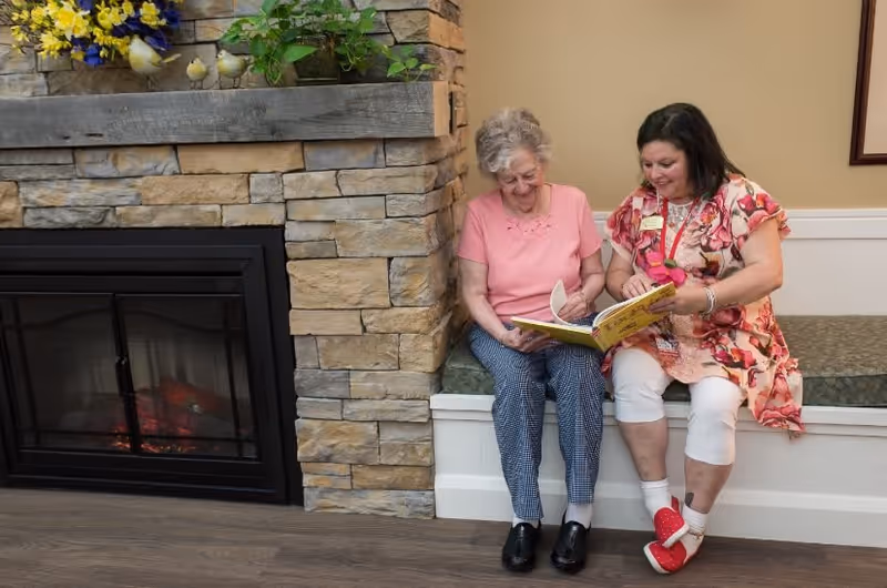 An elderly woman and a caregiver sitting together on a cushioned bench next to a stone fireplace. The elderly woman is wearing a pink shirt and checkered pants, while the caregiver is dressed in a floral top and white pants. They are looking at a book and smiling.