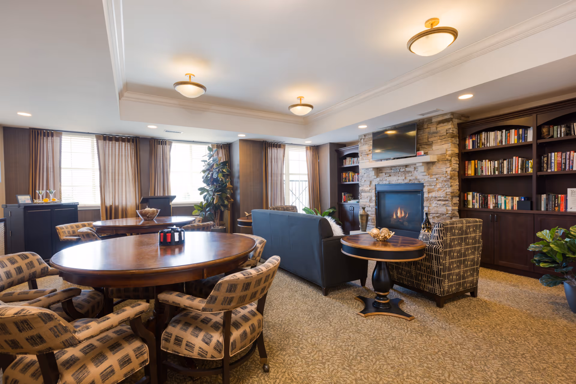 A cozy living room area in an assisted living facility featuring a stone fireplace with a TV mounted above it, built-in bookshelves filled with books, a blue sofa, patterned armchairs, round wooden tables, large windows with brown curtains, and warm ceiling lights.
