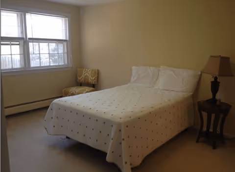 A simple bedroom with a bed covered in a white bedspread with small patterns, two white pillows, a wooden side table with a lamp, a patterned chair near a window with blinds, and beige walls.