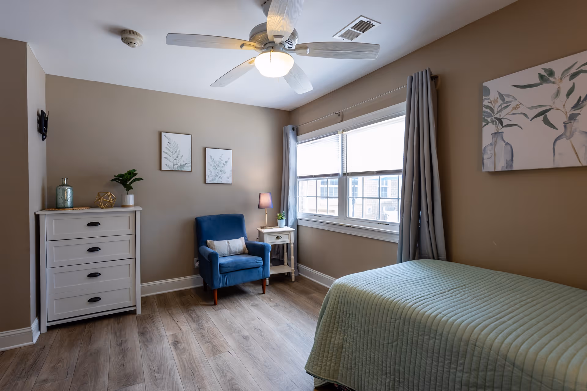 A furnished bedroom with a single bed, blue armchair, white dresser, nightstand, and a large window.