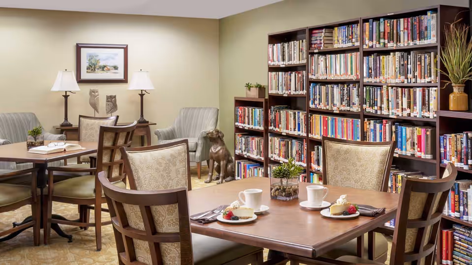 A cozy library room with wooden bookshelves filled with books along the wall. There are two tables with chairs around them, one table has two plates with slices of cake and two cups. The room also features two armchairs, two table lamps, a framed painting on the wall, and a decorative dog statue on the floor.