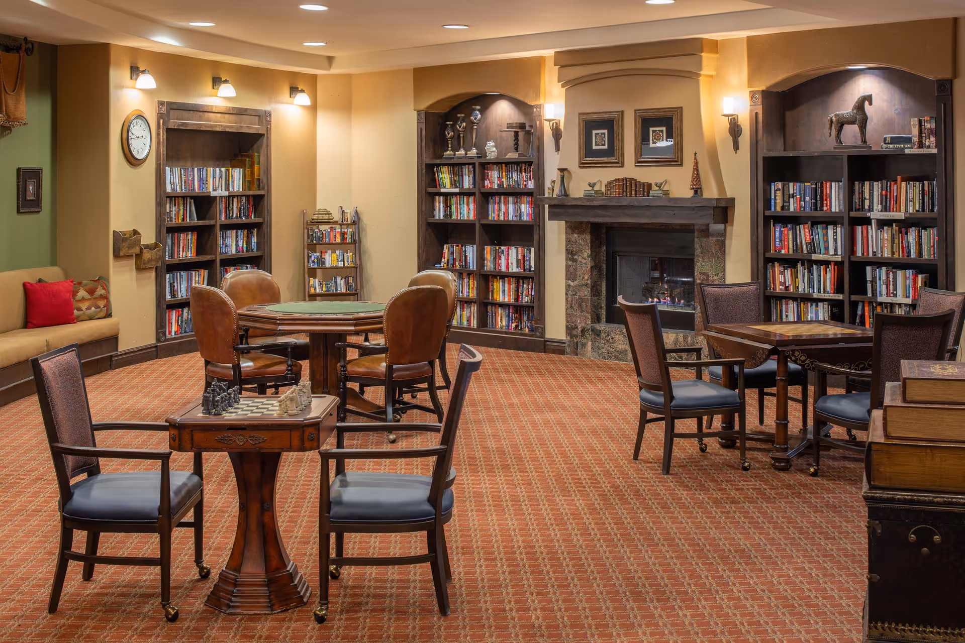 A cozy common area with multiple tables and chairs arranged for games and reading. There are bookshelves filled with books along the walls, a fireplace in the center, and a chessboard on one of the tables. The room has warm lighting and a patterned carpet.