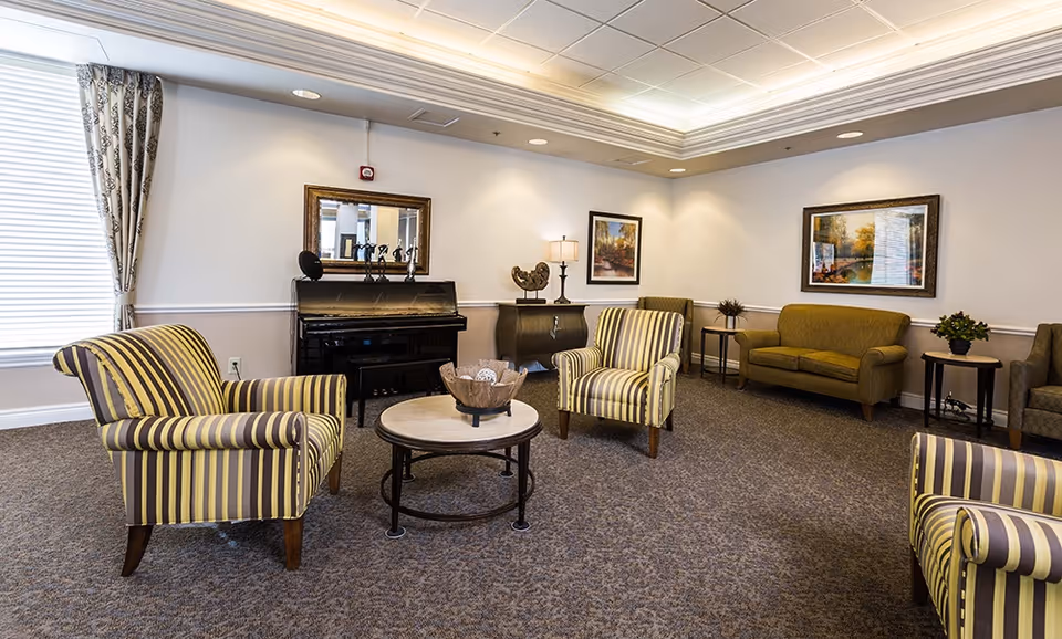 A bright senior living lounge with striped armchairs, sofas, a coffee table and a piano against the wall.