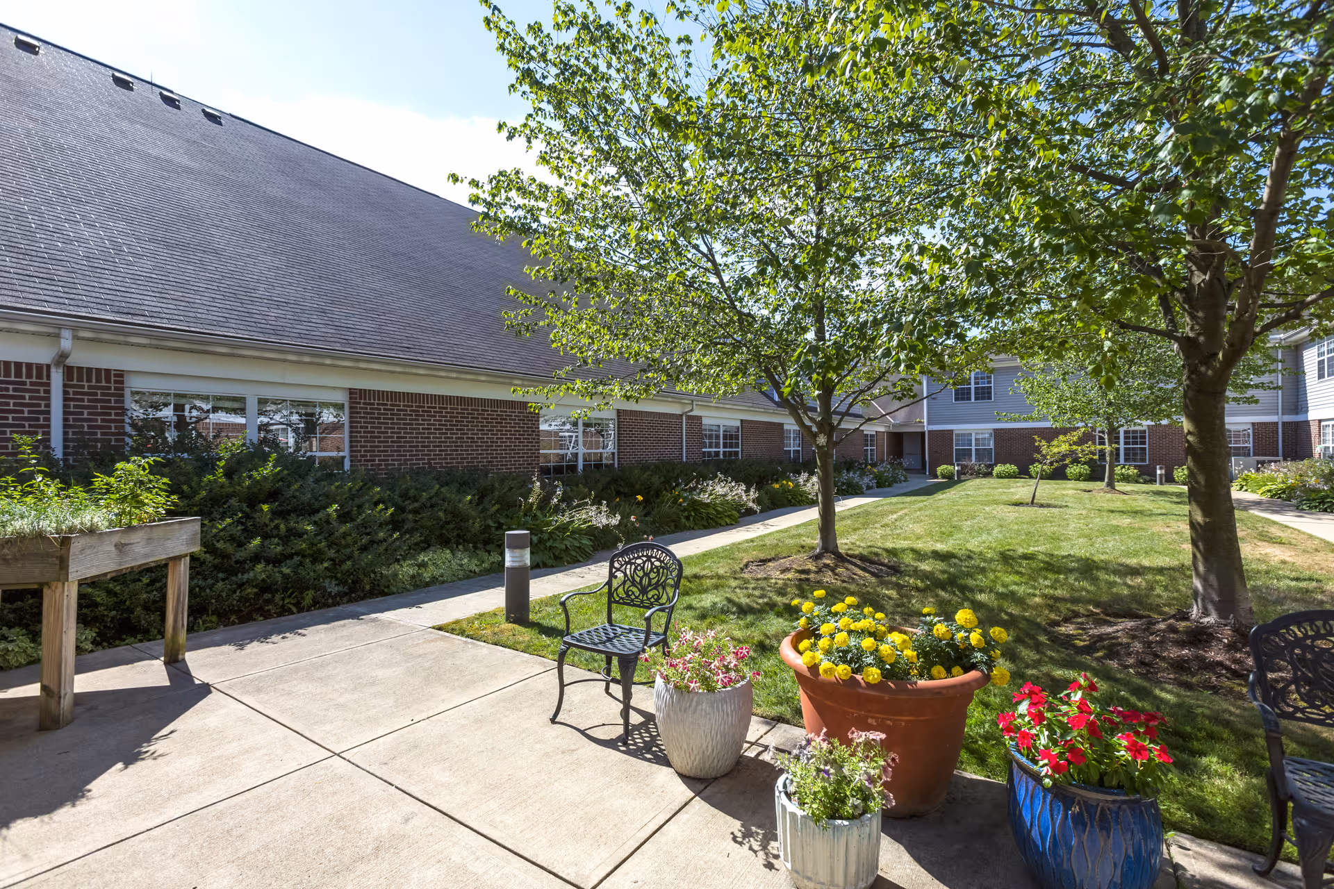 Outdoor courtyard area at Independence Village of Avon Lake featuring a paved walkway, green grass, trees, and several potted flowers. There are black metal chairs placed along the walkway and a raised wooden planter box on the left side. The building with brick and siding is visible in the background under a clear blue sky.