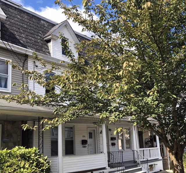 Gray townhouse-style building with a front porch, steps, and a large tree obscuring facade.
