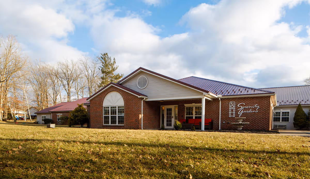 Exterior view of a single-story brick building with a red metal roof and a covered entrance. The building has large windows and a sign that reads 'Serenity Gardens' on the front wall. The surrounding area includes a grassy lawn and leafless trees under a partly cloudy sky.