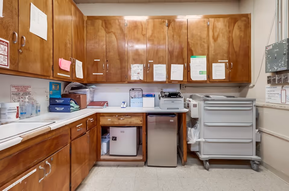 A small utility room with wooden cabinets mounted on the walls and below the countertop. The countertop holds various office supplies, a printer, and some paperwork. Below the counter, there is a small refrigerator and a mini fridge. On the right side, there is a gray rolling cart with drawers and a wall-mounted electrical panel with a clock-in sheet nearby. The floor is tiled, and the room appears to be well-lit.