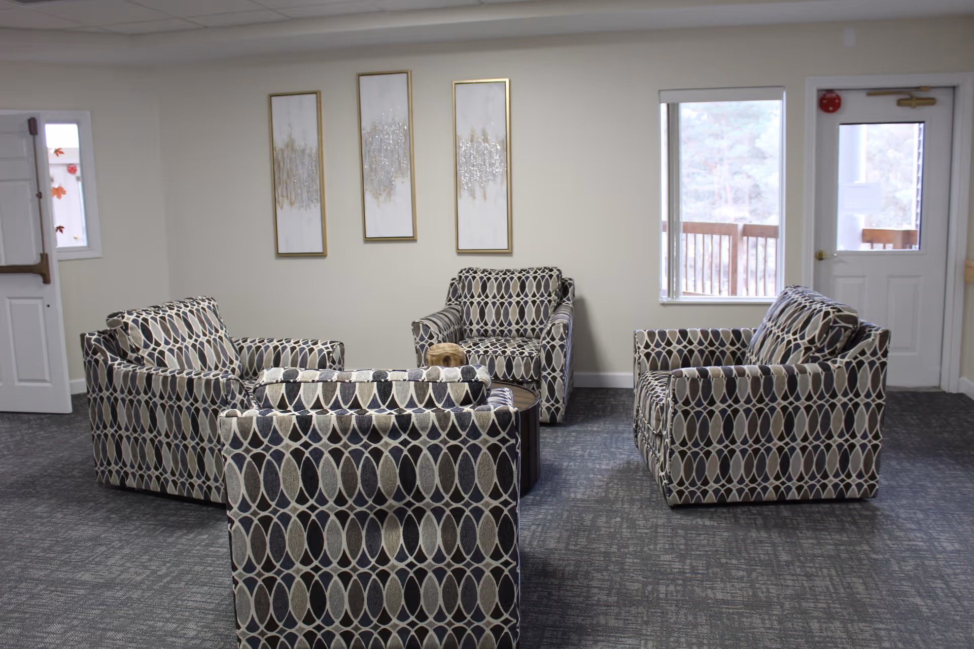 A seating area in a senior living facility with four patterned armchairs arranged around a small round table. The room has light-colored walls with three vertical abstract paintings and two windows, one next to a door leading outside. The floor is carpeted in a dark gray pattern.