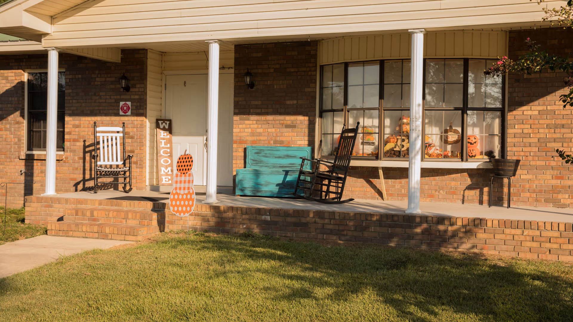 Front porch of a brick building with white columns, two rocking chairs, a turquoise bench, and autumn-themed decorations including a welcome sign and pumpkin cutouts. The porch overlooks a green lawn.