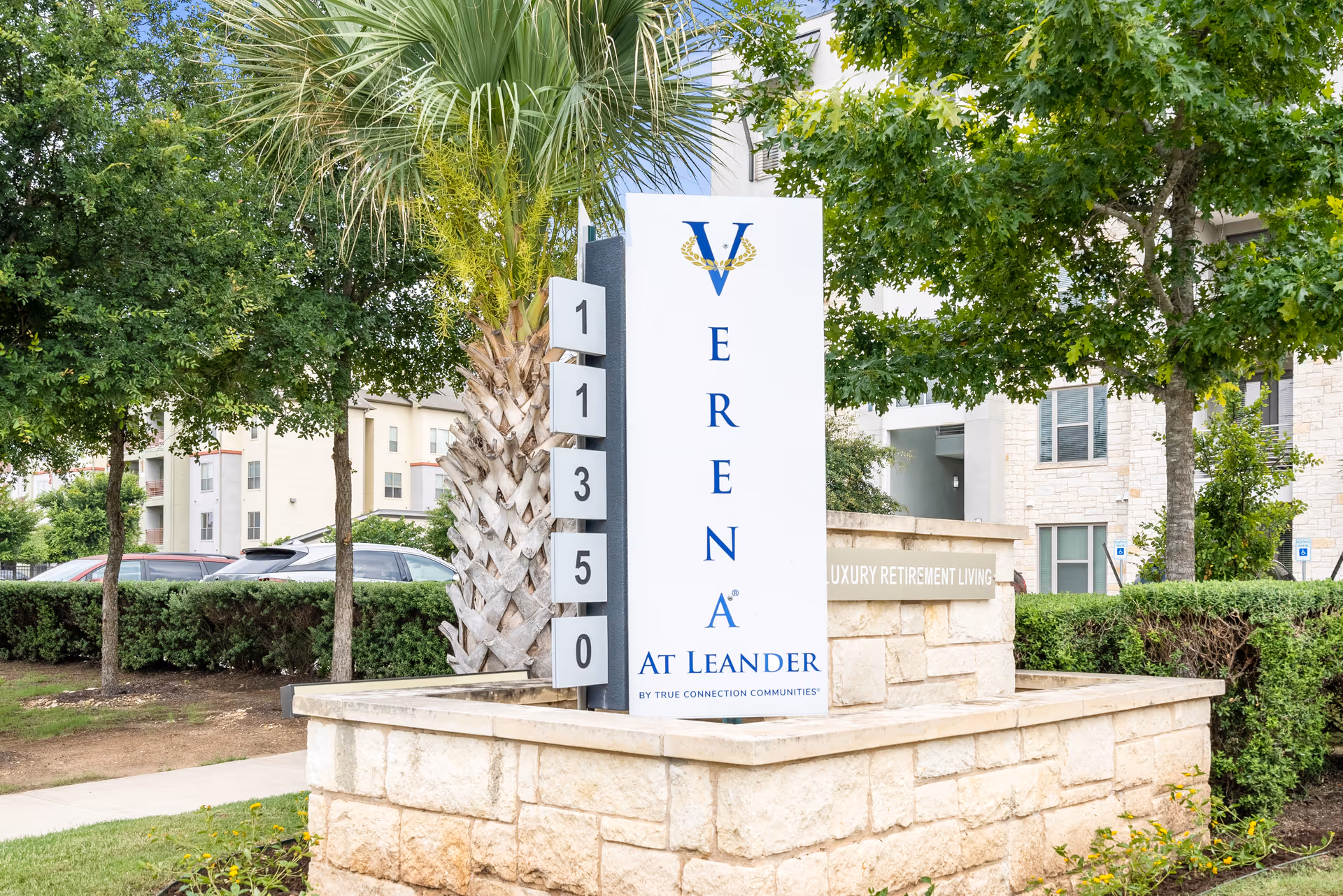 Outdoor view of the entrance sign for Verena at Leander, a luxury retirement living community, surrounded by trees, bushes, and parked cars.