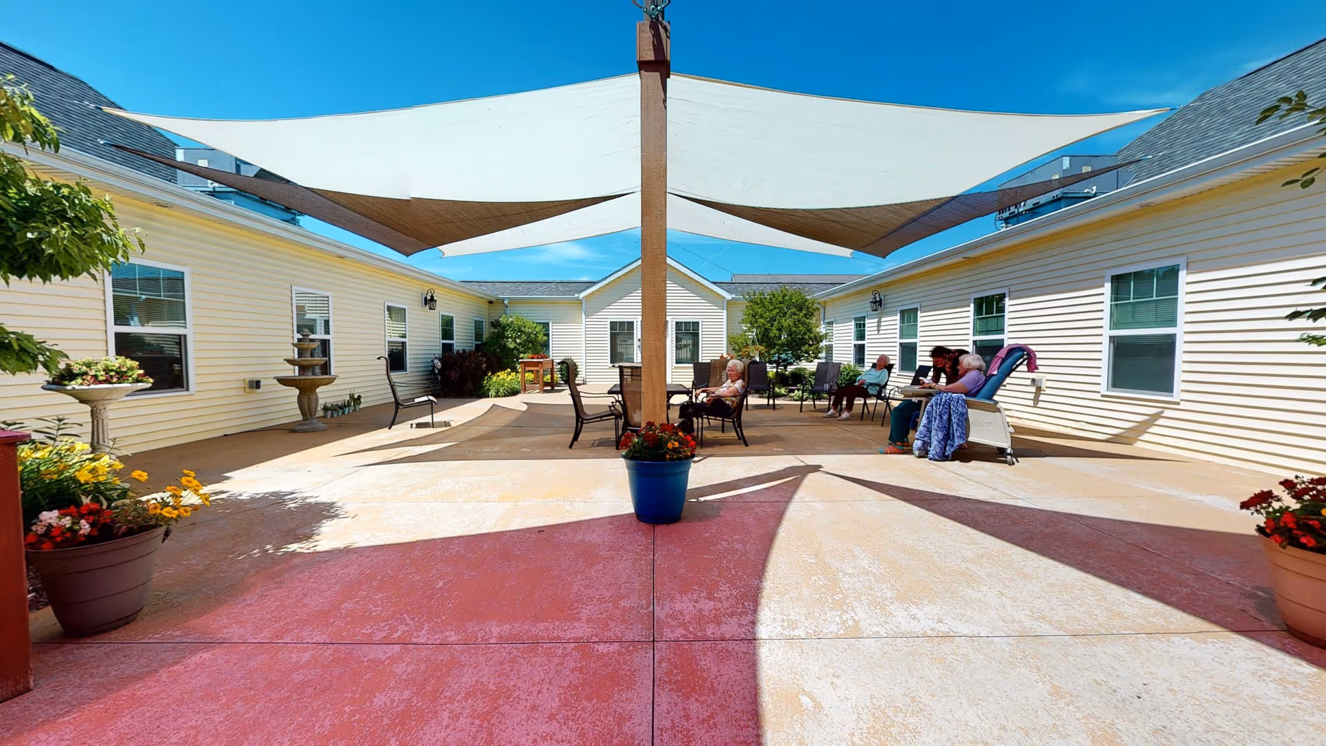 Outdoor courtyard area of Vicinia Gardens Luxury Retirement Living - Assisted Living with several elderly residents sitting on chairs under shade sails. The courtyard is surrounded by single-story buildings with light-colored siding and windows. There are potted plants and flowers around the space, and a clear blue sky overhead.