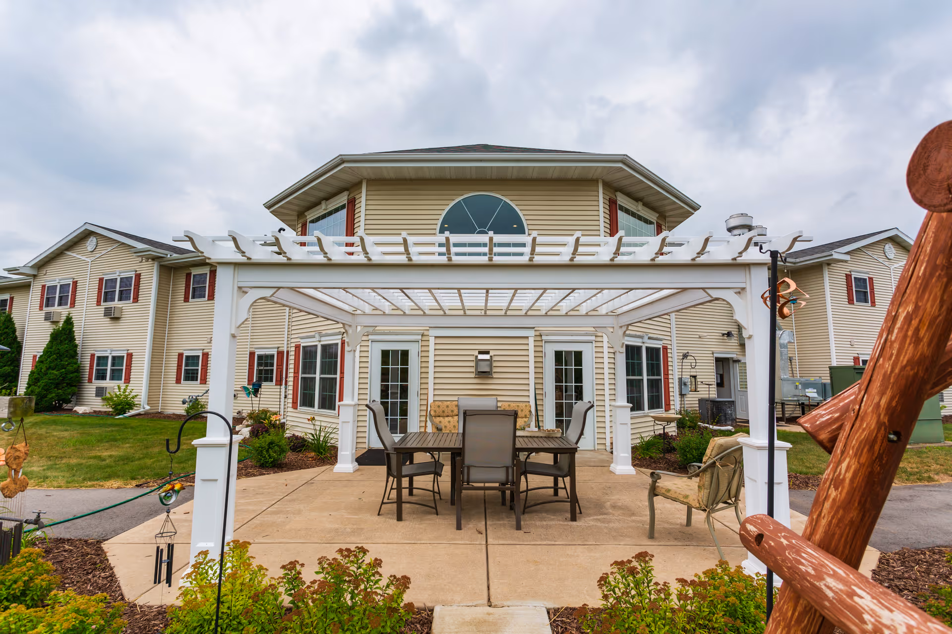 Outdoor patio area at The Cottages at Meadowlands Senior Living featuring a white pergola with a table and six chairs underneath, surrounded by greenery and a beige two-story building with red shutters in the background under a cloudy sky.