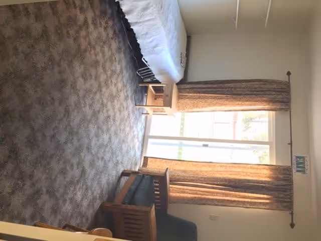 A small bedroom with a single bed covered in a white blanket, a wooden nightstand next to the bed, a window with brown curtains letting in natural light, and a wooden chair with green upholstery. The floor is carpeted with a patterned design.