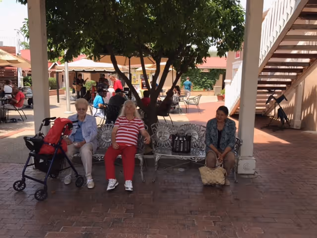 Three people seated on a decorative metal bench under a shaded tree in a courtyard with tables, umbrellas and a nearby staircase.