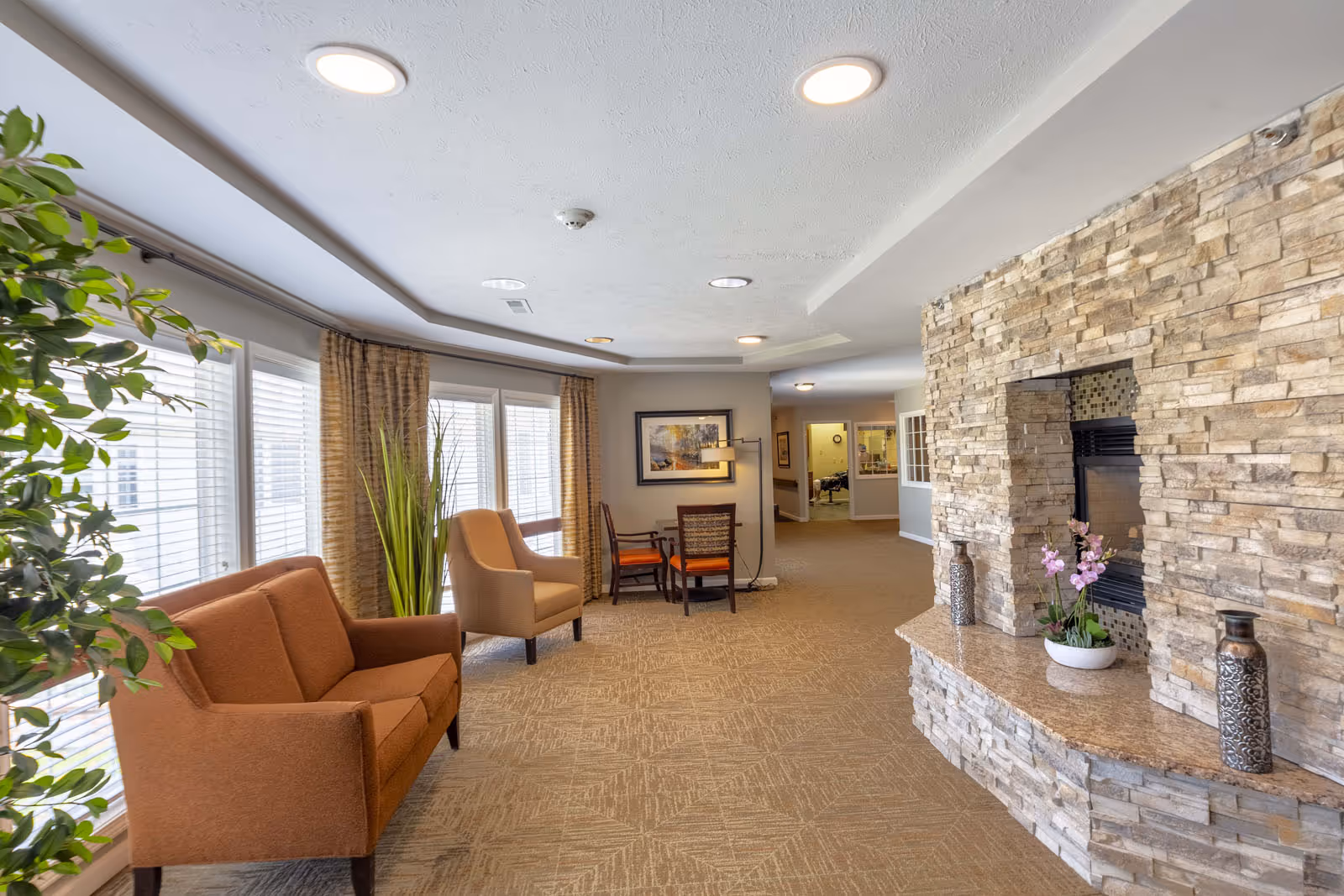 A bright communal lounge with upholstered chairs, large windows, plants, and a stone fireplace.