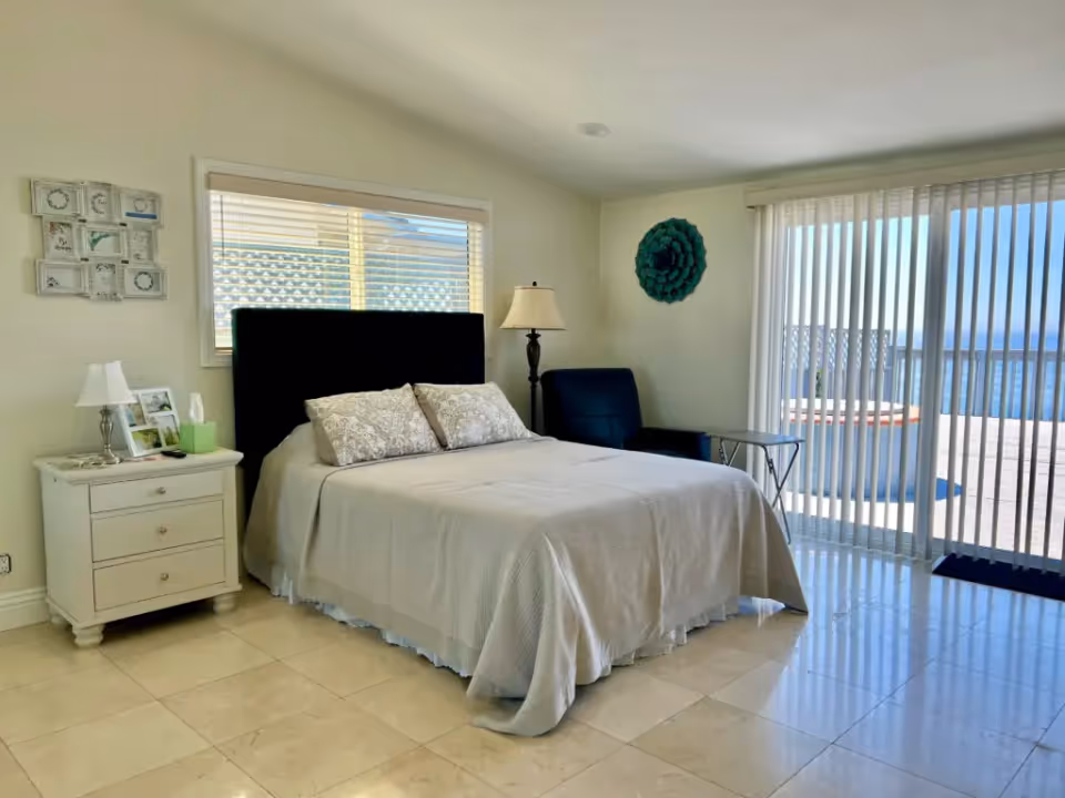 A bright bedroom with a neatly made bed featuring a black headboard and two patterned pillows. To the left of the bed is a white nightstand with a lamp, tissue box, and framed photos. A window with blinds is above the bed. To the right, there is a black armchair, a floor lamp, and a small glass side table. Large sliding glass doors with vertical blinds open to an outdoor patio with a view of the ocean.