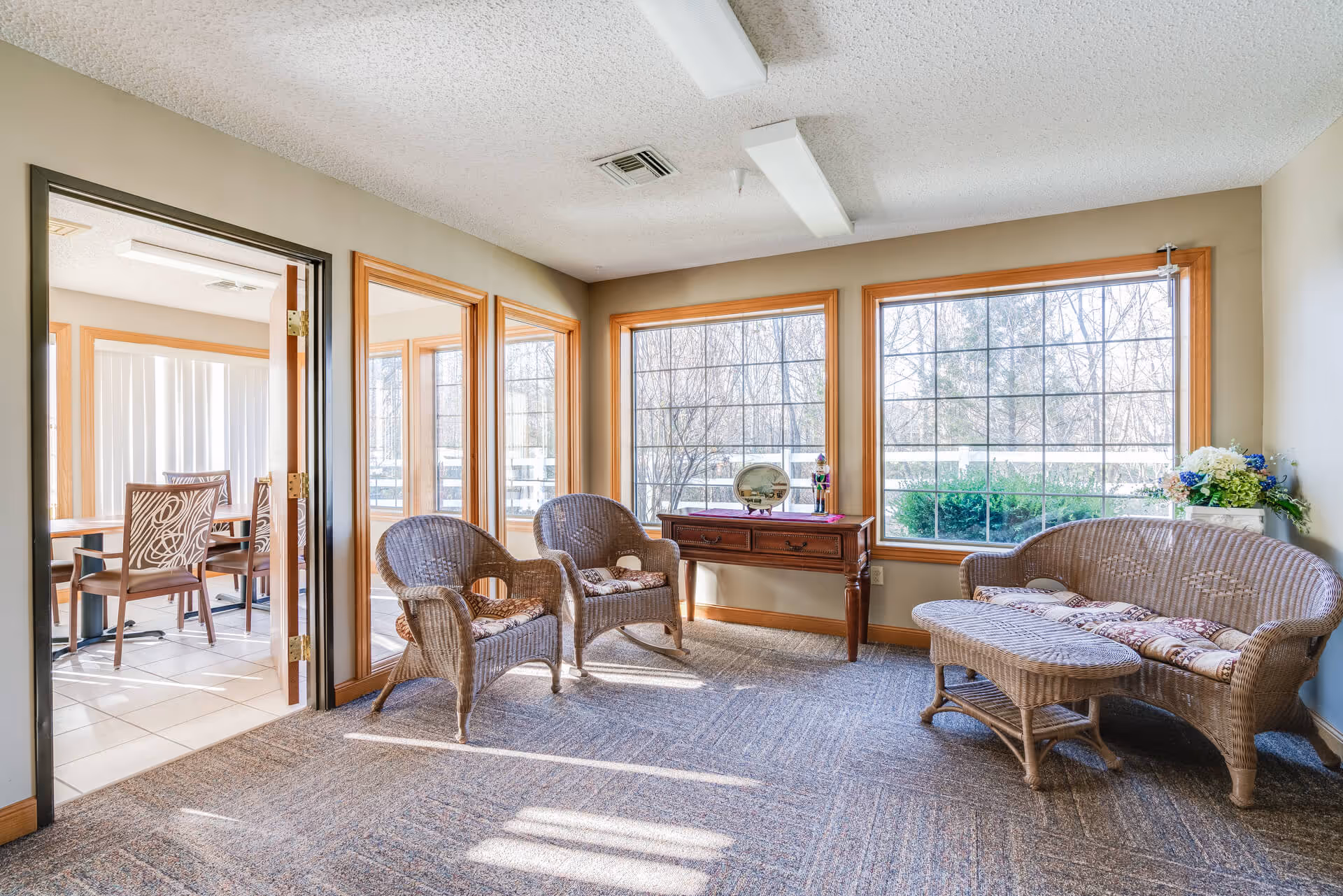 A bright sitting area in a senior living facility with large windows letting in natural light. The room features two wicker armchairs, a wicker loveseat with cushions, a matching wicker coffee table, and a wooden console table with decorative items. Adjacent to this area is a dining room with a table and chairs visible through an open doorway.