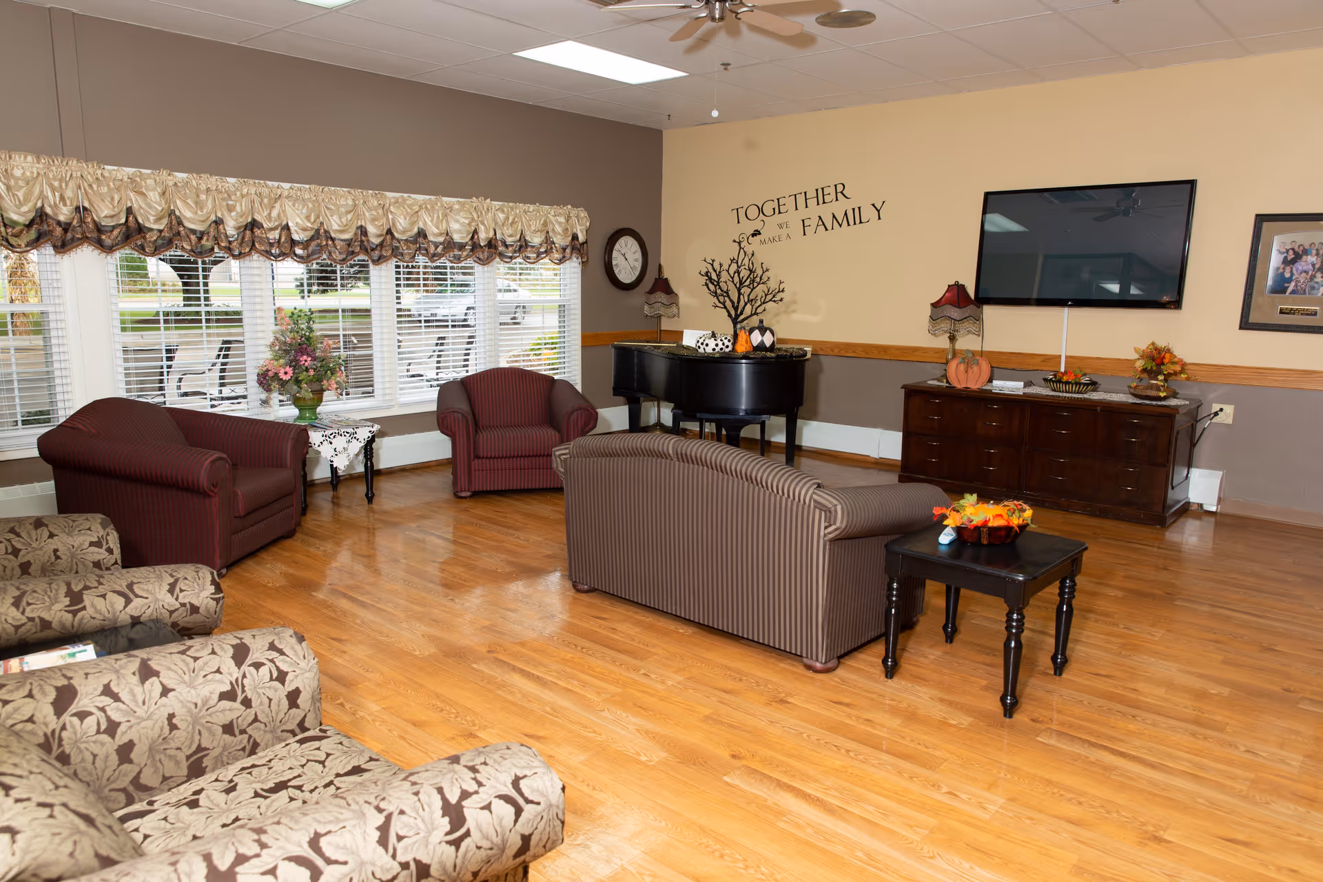 A cozy living room with wooden flooring, featuring a striped sofa, two patterned armchairs, and two maroon armchairs. There is a small black side table with a floral arrangement, a black grand piano with decorative items on top, and a wooden cabinet with a flat-screen TV mounted above it. The wall has a decal that reads 'Together we make a family.' Large windows with valances let in natural light.