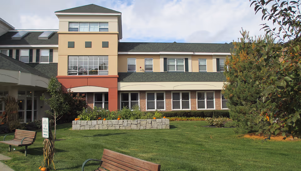 Exterior view of Hearthstone Alzheimer Care at Marlborough, showing a two-story building with beige and red accents, multiple windows, a green lawn with benches, small trees, and a stone planter with pumpkins.