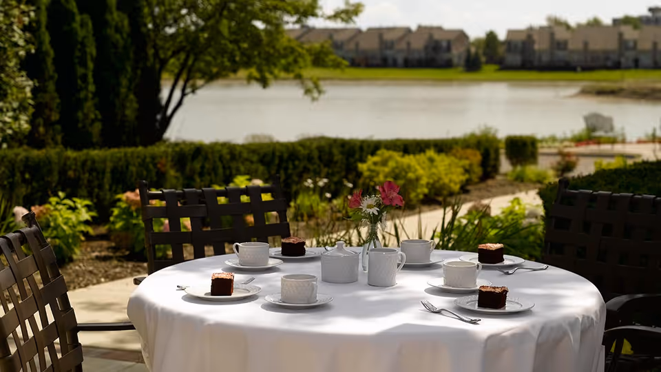 Outdoor round table covered with a white tablecloth set with white teacups, saucers, a teapot, a creamer, and plates each holding a piece of chocolate cake. The table is surrounded by dark metal chairs with woven backs. In the background, there is a garden with green shrubs and trees, a walking path, and a body of water with buildings visible across the water.