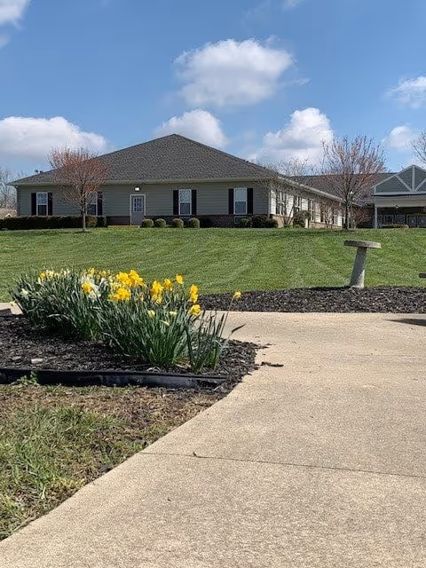 A single-story building with a gray roof and light-colored walls, surrounded by a well-maintained lawn and flower beds with yellow daffodils under a blue sky with scattered clouds.