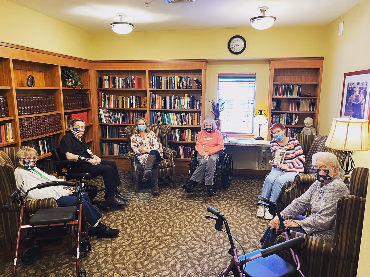A group of six elderly individuals wearing face masks sitting in a cozy room with bookshelves filled with books. The room has patterned carpet, wooden bookshelves, a window with blinds, a clock on the wall, and a floor lamp. Some individuals are seated in armchairs while others use wheelchairs or walkers.