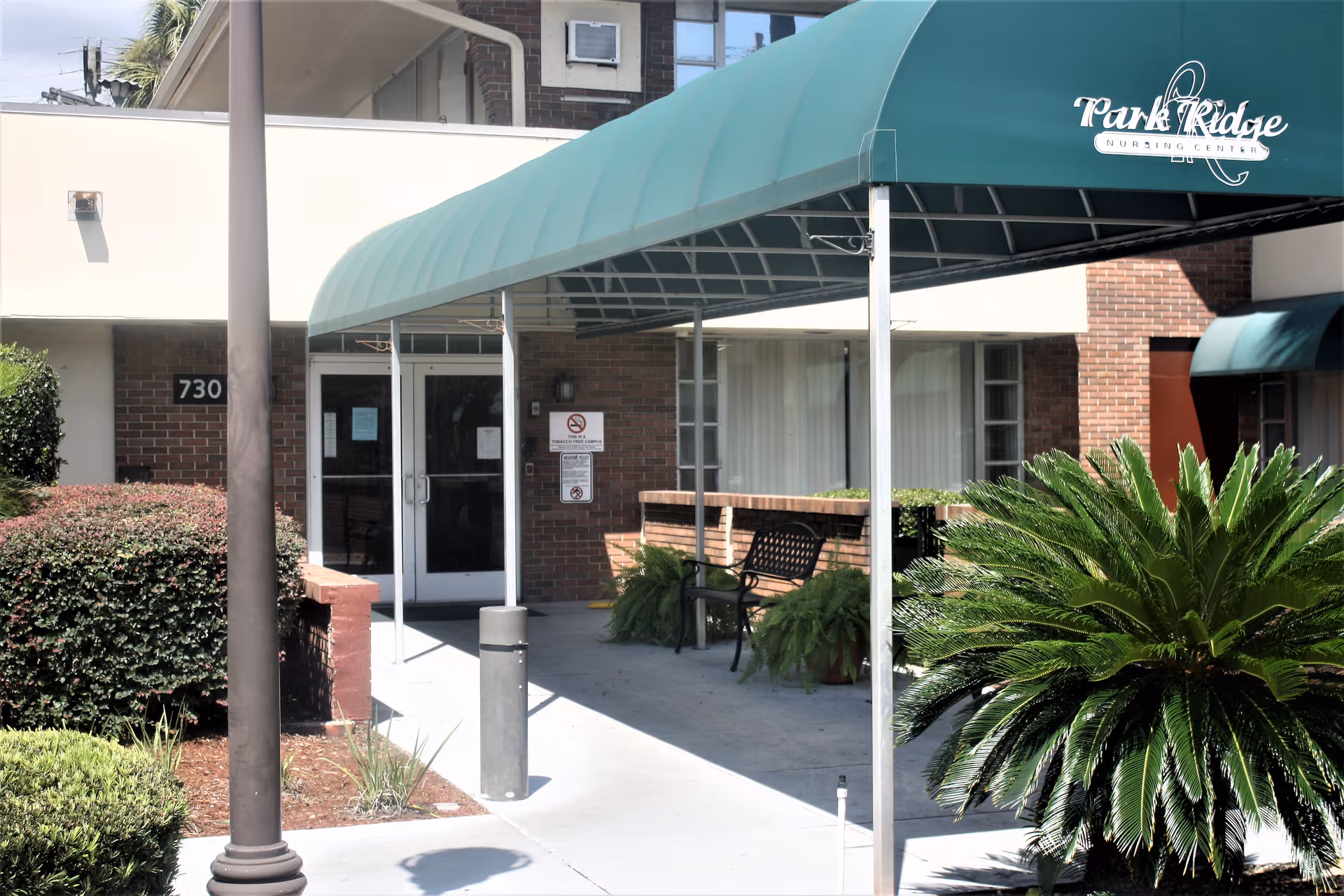 Entrance to Park Ridge Nursing Center with a green awning over the walkway, a bench, and surrounding greenery including bushes and a palm-like plant.