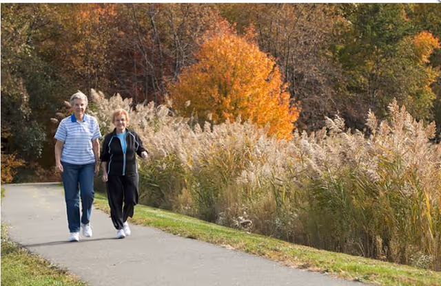 Two elderly women walking together on a paved path surrounded by tall grasses and autumn-colored trees in the background.
