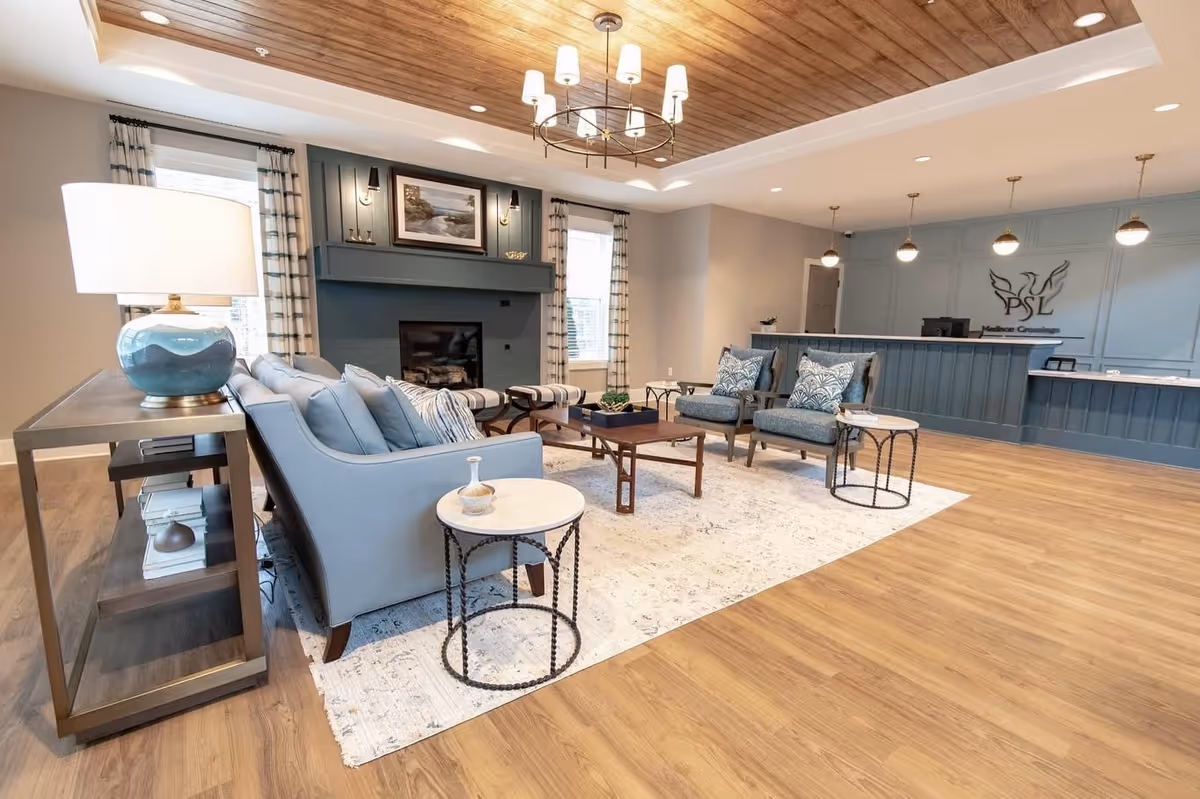 A spacious and well-lit living room area with a light blue sofa, two matching armchairs with patterned cushions, a wooden coffee table, and two round side tables with marble tops. The room features a fireplace with a painting above it, two windows with curtains, a wooden ceiling accent with a chandelier, and a reception desk in the background with pendant lights and a logo on the wall.