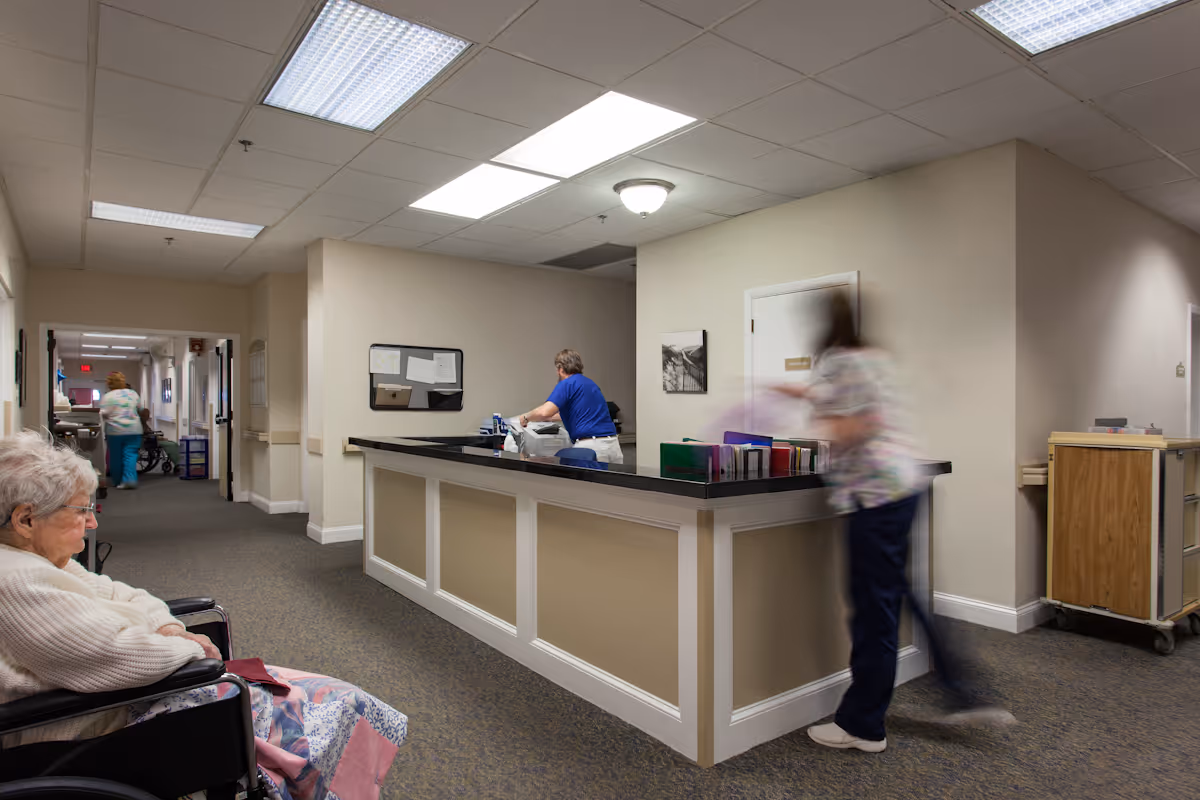 Interior view of a nursing center hallway with a reception desk. An elderly woman in a wheelchair is seated on the left side, while two staff members are behind and near the reception desk. The hallway extends into the background with another staff member walking away.