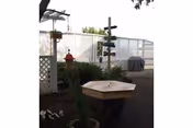 Small fenced outdoor patio with potted plants, a wooden hexagonal table, a decorative signpost, and a covered grill.