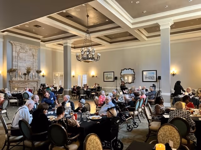 A large dining room filled with elderly people seated at round tables covered with black tablecloths, eating and socializing. The room has high ceilings with decorative molding, a chandelier, wall sconces, framed artwork, and a large ornate fireplace. Some people use walkers and wheelchairs.