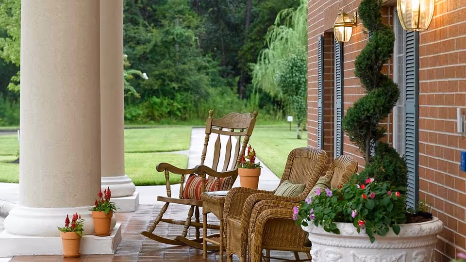 A porch area with a wooden rocking chair and two wicker chairs with cushions, surrounded by potted plants and flowers. The porch has large columns and a brick wall with outdoor lanterns, overlooking a green lawn and trees in the background.