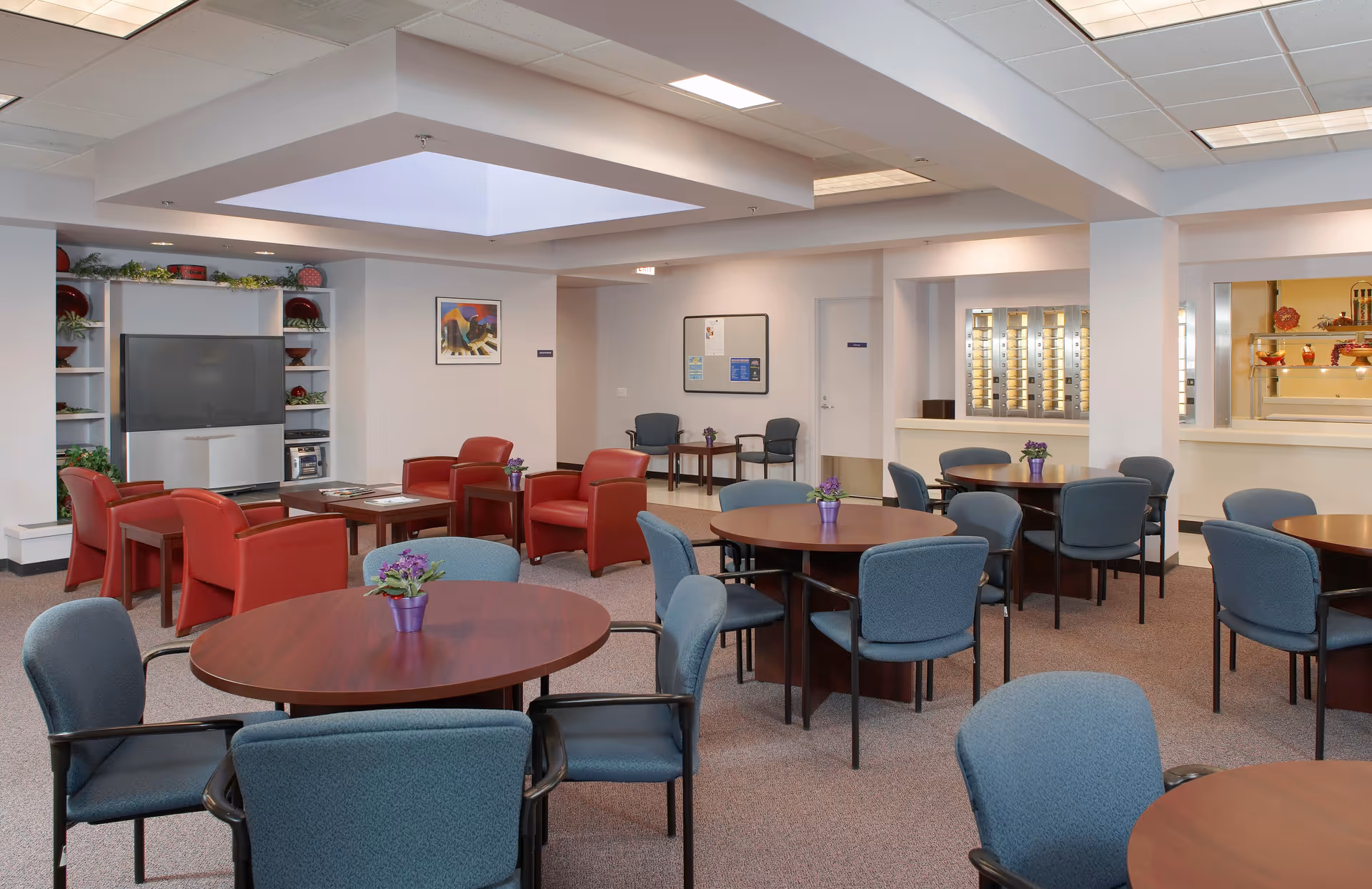 A spacious common area in a senior living facility featuring multiple round wooden tables with blue cushioned chairs arranged around them. In the background, there is a seating area with red armchairs facing a large television set on a white entertainment unit with shelves decorated with plants and red plates. The room has a neutral color palette with beige walls and carpeted floors, and a skylight in the ceiling providing natural light. Small purple flower pots are placed on each table.