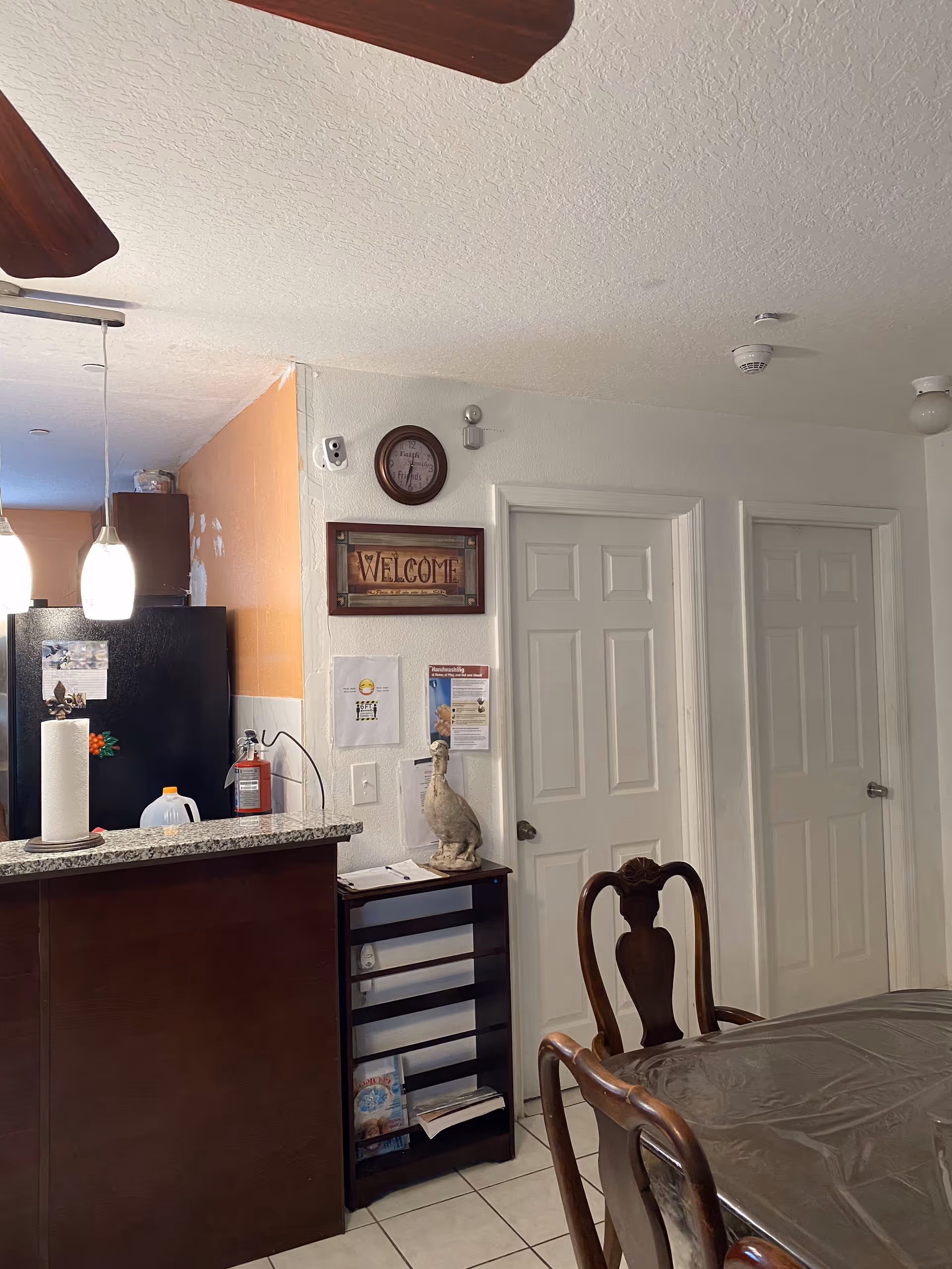 Dining area and kitchen pass-through with a table and chairs, countertop with paper towel and jug, refrigerator, wall clock and a framed "WELCOME" sign.