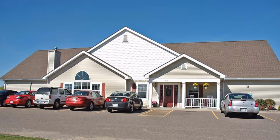 Single-story beige senior living building with a front porch and several cars parked in front under a clear blue sky.