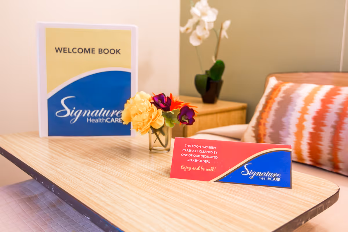 A wooden table with a small vase of colorful flowers, a welcome book with the Signature HealthCARE logo, and a card stating the room has been carefully cleaned. In the background, there is a chair with a patterned cushion and a small plant on a side table.