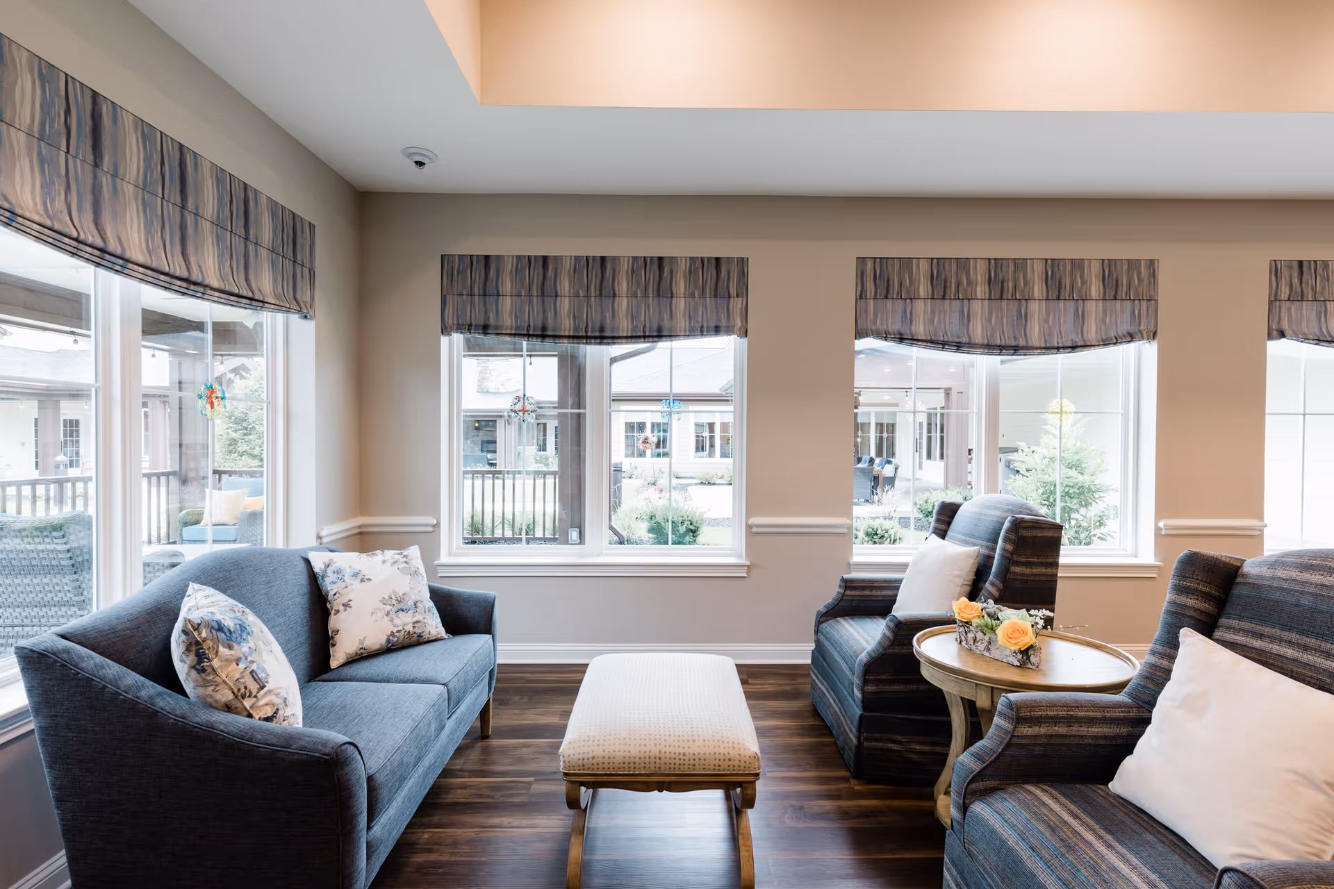 A bright and cozy living room area with a blue sofa adorned with floral pillows, two striped armchairs with white pillows, a small round wooden table with a flower arrangement, and a cushioned bench in the center. Large windows with patterned valances allow natural light to fill the room and offer a view of the outdoor courtyard.