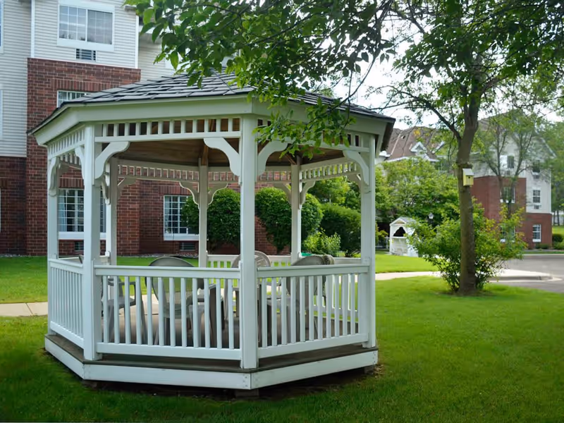 A white wooden gazebo with a shingled roof situated on a green lawn in front of a brick and white siding building. Inside the gazebo are several white chairs arranged around a table. Trees and bushes surround the area, creating a peaceful outdoor setting.