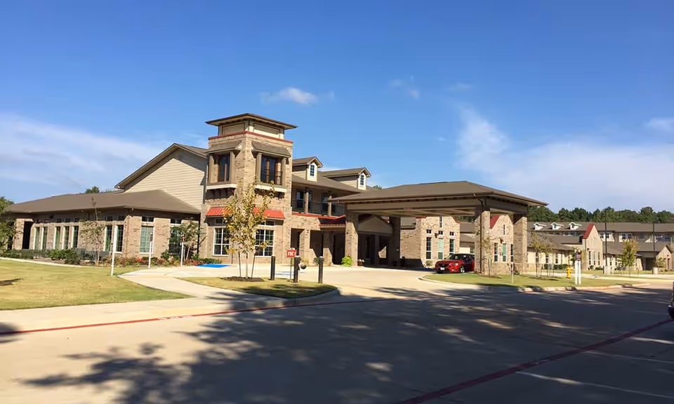 Front exterior of a brick senior living building with a covered entrance/porte-cochere and landscaped lawn under a clear blue sky.