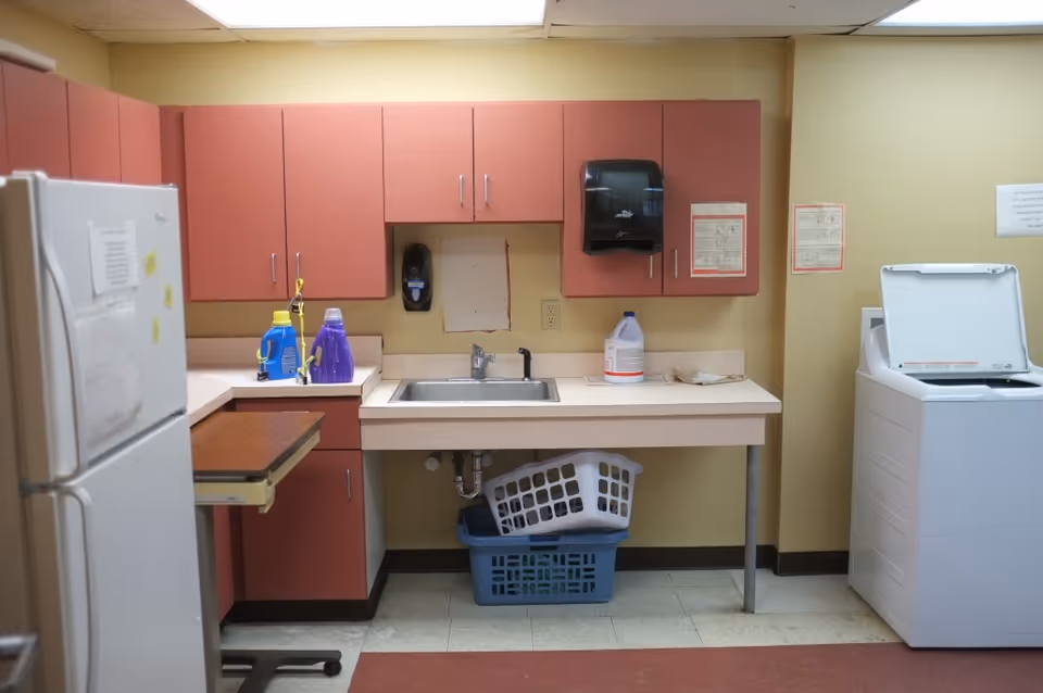 Small kitchenette/laundry room with pink cabinets, a sink, detergent bottles, a washing machine and a refrigerator.